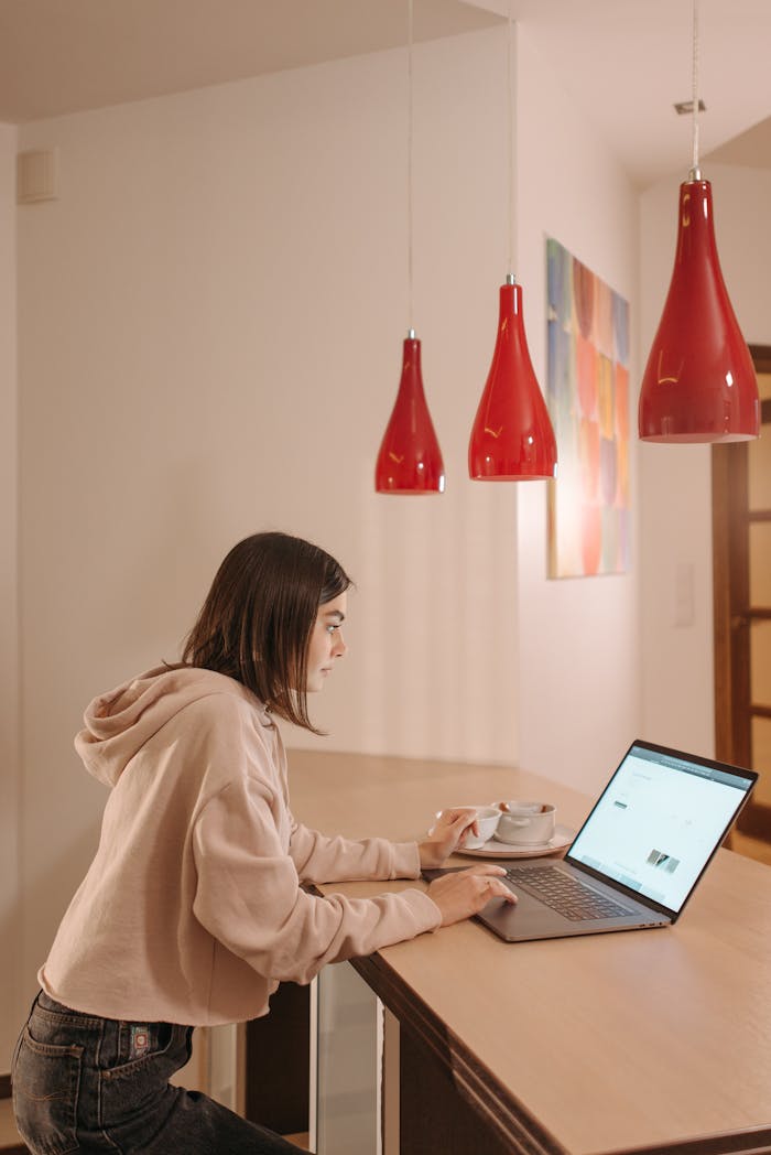 A woman in a casual hoodie shops online from her laptop in a modern kitchen with red pendant lights.
